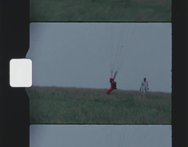 A parachutist holds the lines as he floats down with their parachute and lands on the ground near a man standing in the field