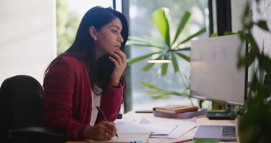 A woman sitting at her desk using her computer then writing on paper in front of her