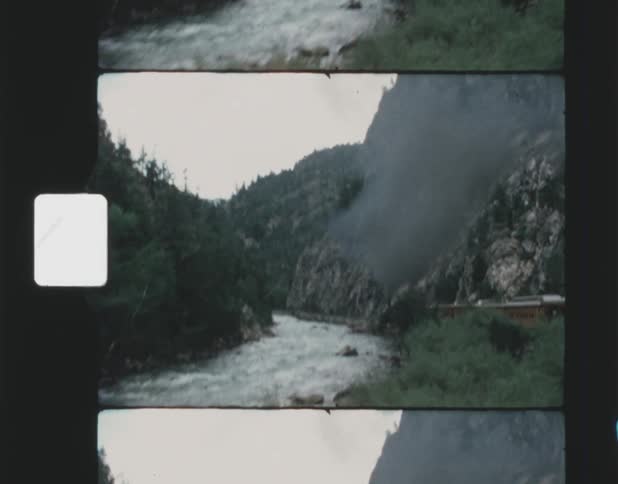 A passenger train travels through a mountainous area with a river and an evergreen forest