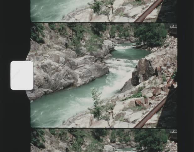 A stream runs through forested mountains along a railway as black smoke rises from the coming train