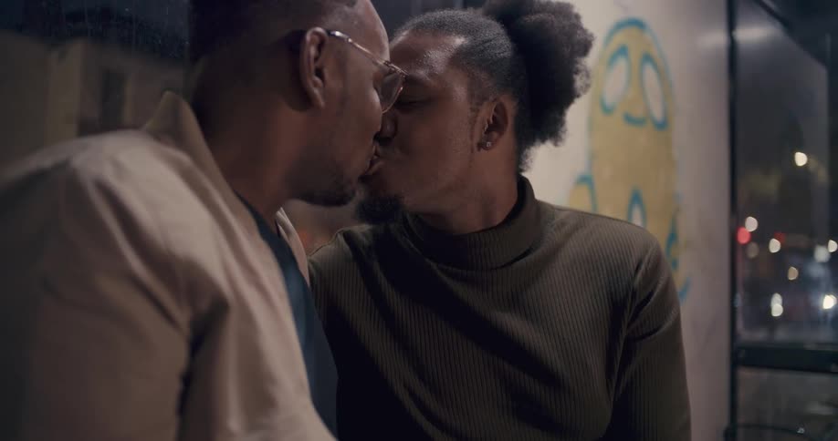 Two men sit smiling at each other and kiss before looking at an engagement ring.