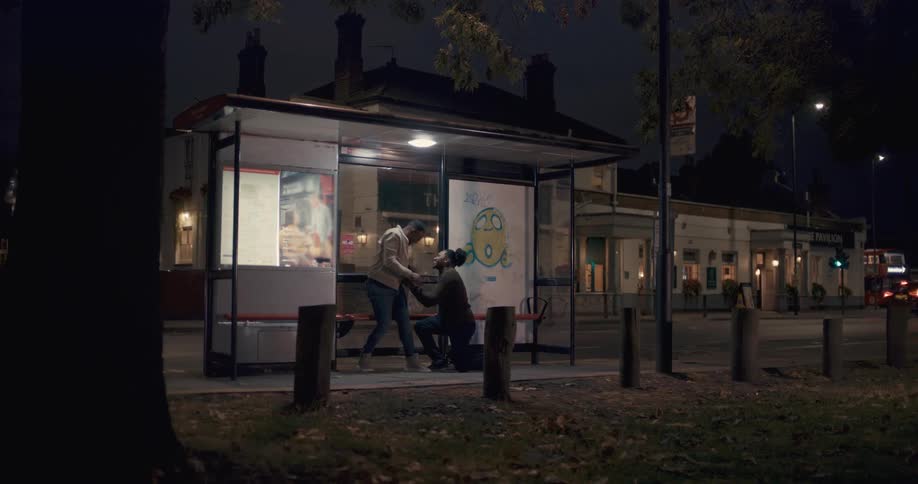 A wide shot of a  man proposing to another man at a bus stop