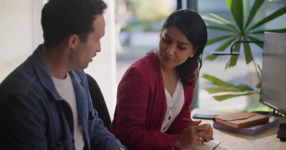 A man takes notes while chatting with a colleague at her desk