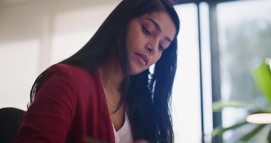 Businesswoman with dark straight hair and red blazer sits at desk and writes down at the office