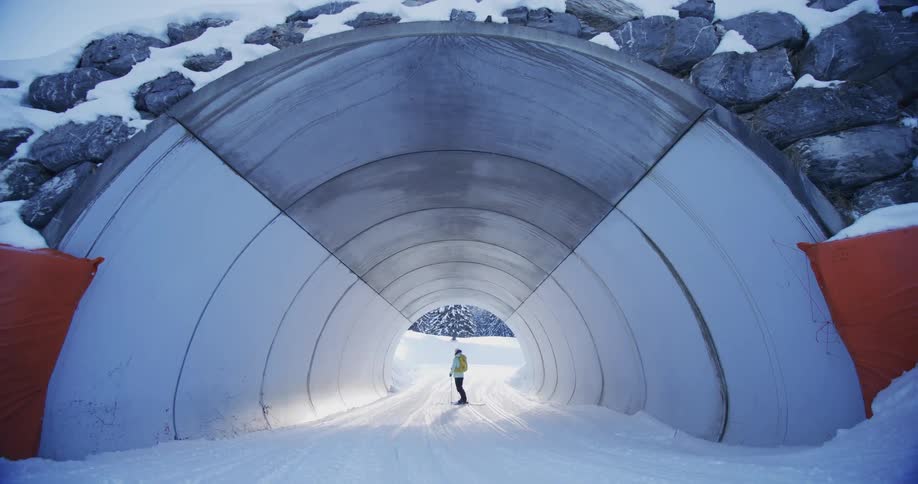 A woman wearing a backpack is standing in the snow in a tunnel holding a ski pole