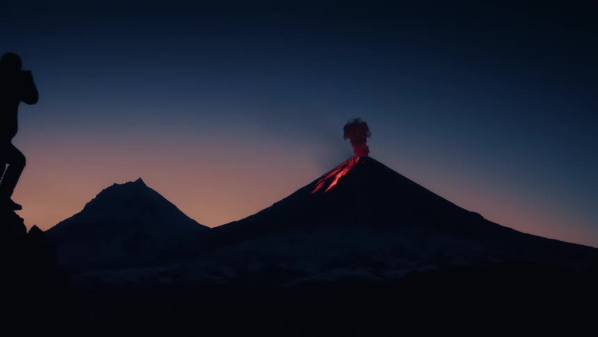 A man is standing with a headlamp and a camera as hot lava pours from a volcano and smoke rises in the darkening sky