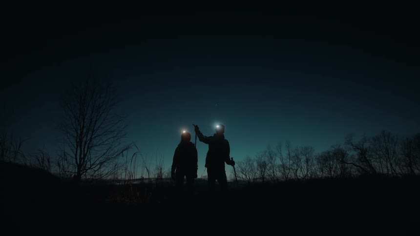 Two people are wearing helmet lights and carrying trekking poles as they walk in the darkness with bare trees behind them