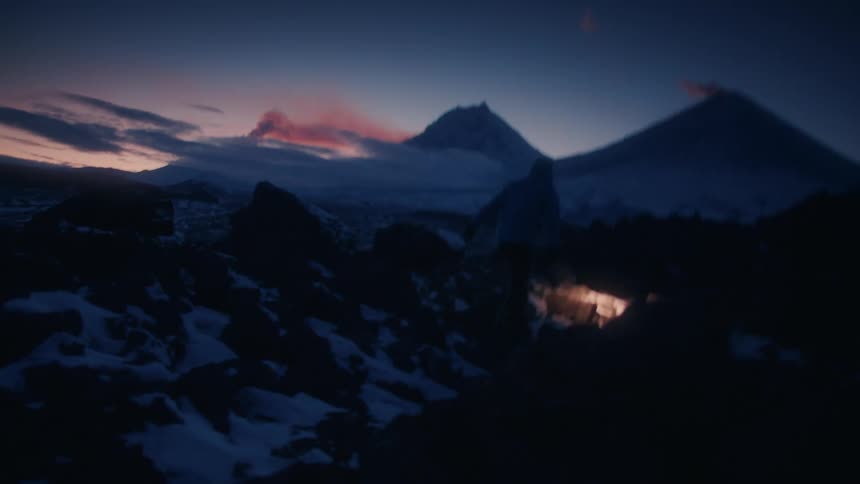 Man walks over snow covered and boulder strewn uneven ground with red clouds hovering over mountains in the background