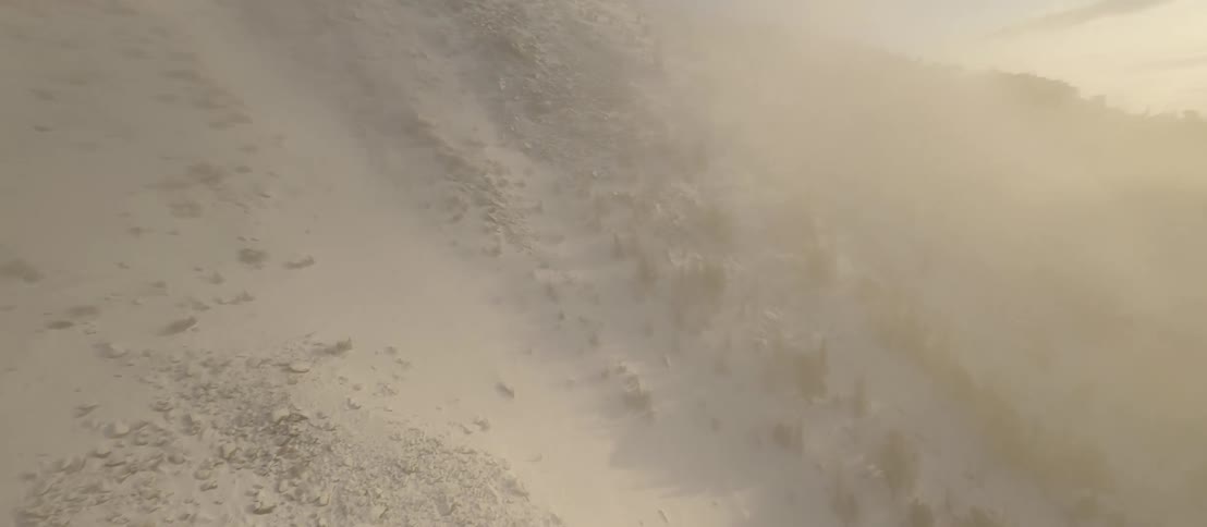The sun shines brightly upon desert mountains with scattered trees and vegetation as a dust storm fills the air