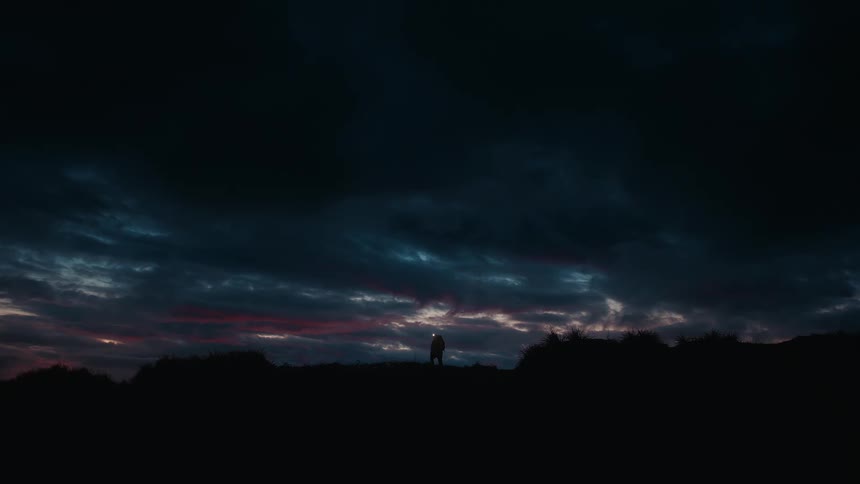 A person hikes up to a mountain ridge under a cloudy nighttime sky
