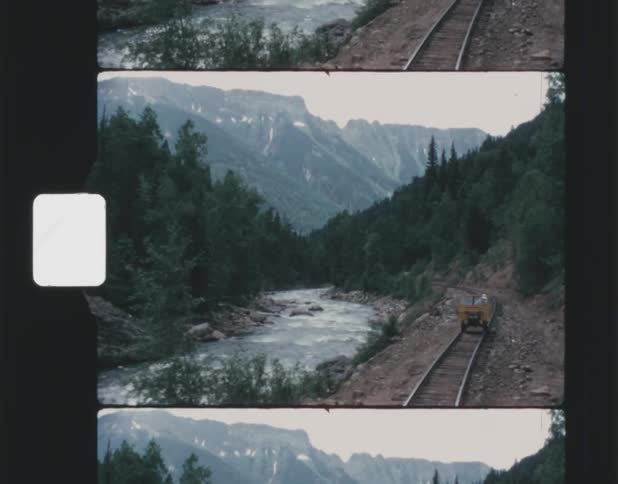 Small carfollows along train track paralleling river at base of forest covered mountain