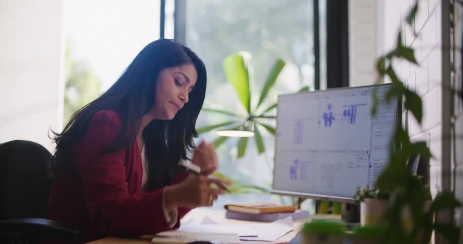 Woman with dark straight hair and red blazer jacket sitting at desk yawns at the office