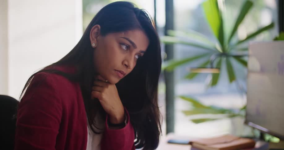 Woman sits in office and makes notes while consulting computer as traffic passes outside window