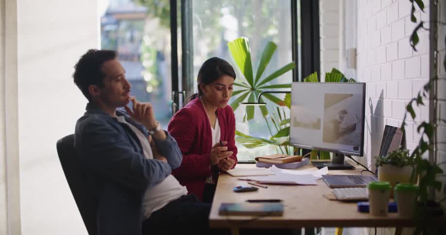 A man and woman sit at a desk together while the woman is writing the man is thinking and the two converse