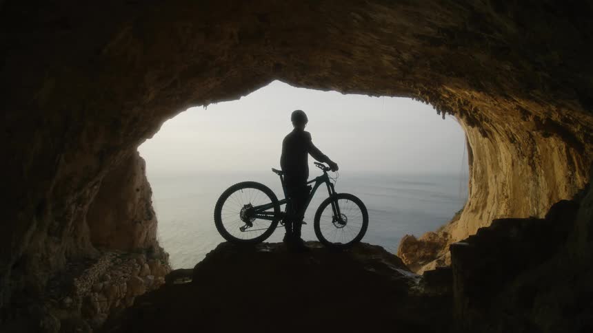 Cyclist with mountain bicycle is silhouetted against the horizon seen from a cave on mountain