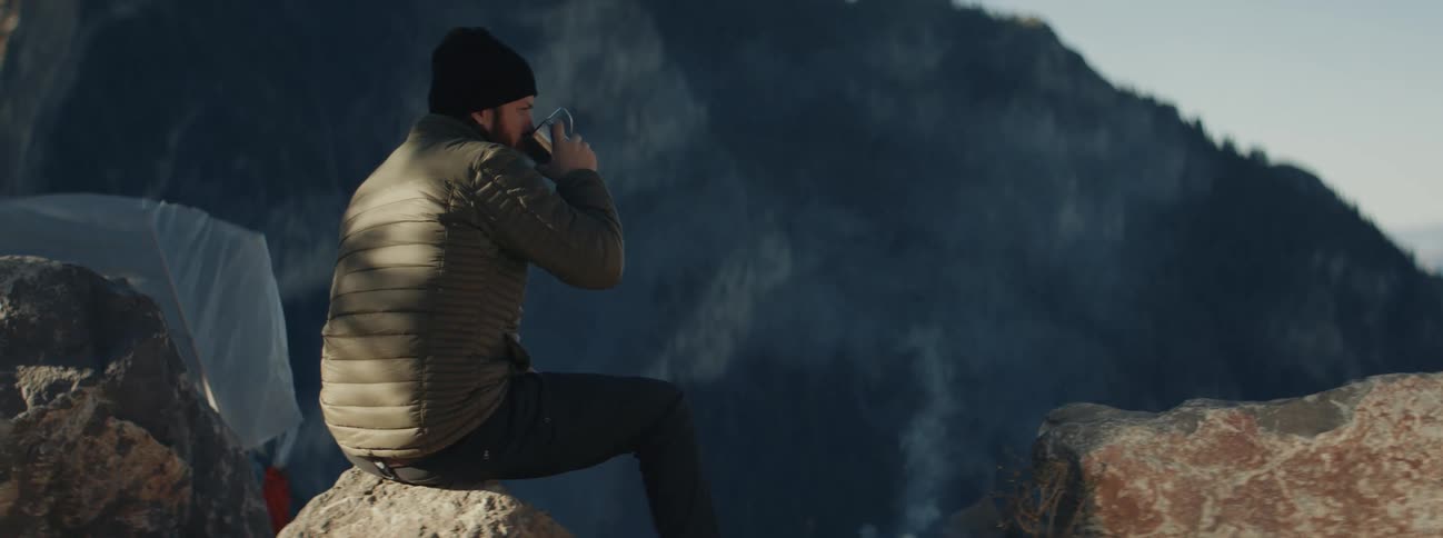 A man drinking coffee next to a campfire in the mountains at sunrise