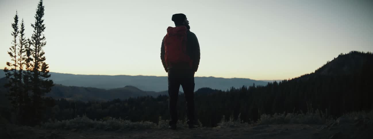 A man standing high in the wilderness looks out over the rolling forested countryside below during sunrise