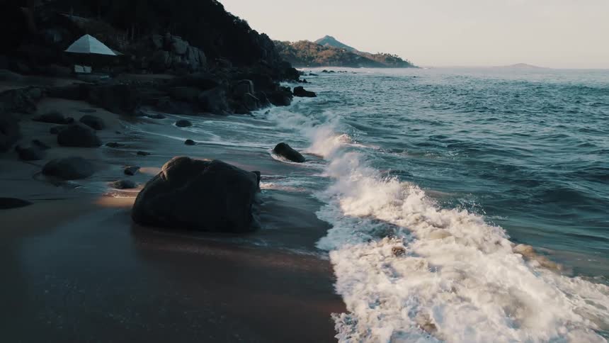 Ocean waves wash into a sandy beach covered in large stones during the day