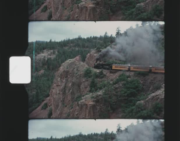 Train releases dark smoke while it moves along cliff on forested mountain