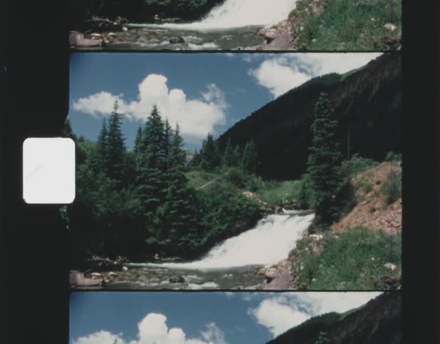A stream flows down a hill near a forest with a mountain in the distance and cumulus clouds in a blue sky