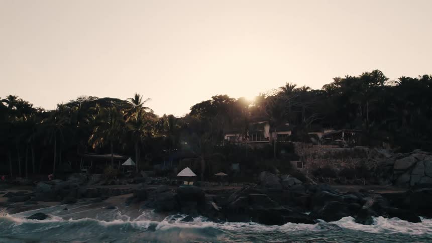 Aerial view of ocean waves breaking against the shore and holiday resort in the jungle at sunset