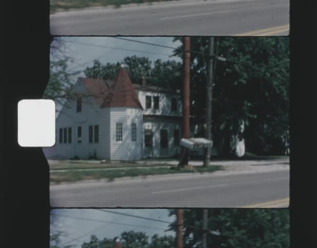 A vintage film shows a neighborhood with houses and trees as someone off camera driving in a car is filming