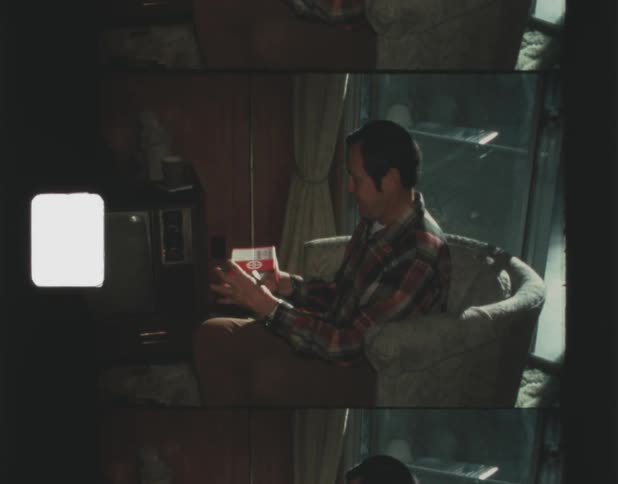Man with red shirt and brown pants sits in armchair in the living room unwrapping a present