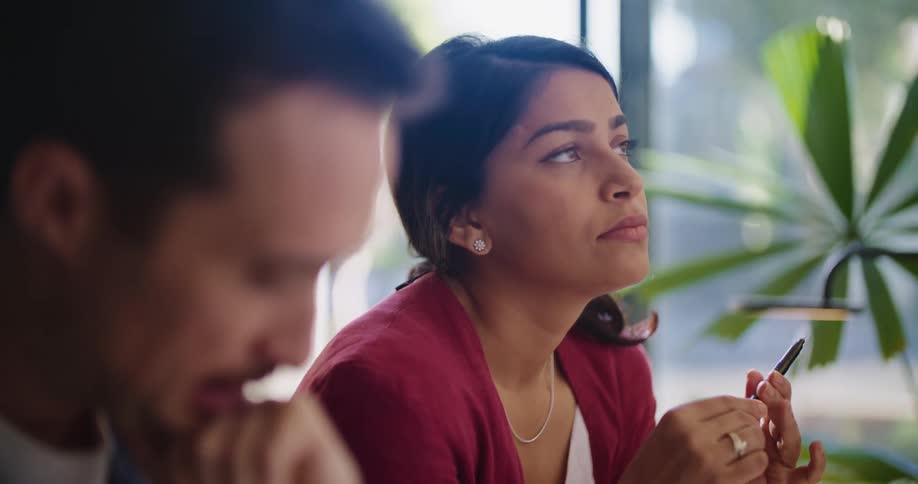 A woman is sitting at a table next to a man as she holds a pen in her hands and looks down at the desk