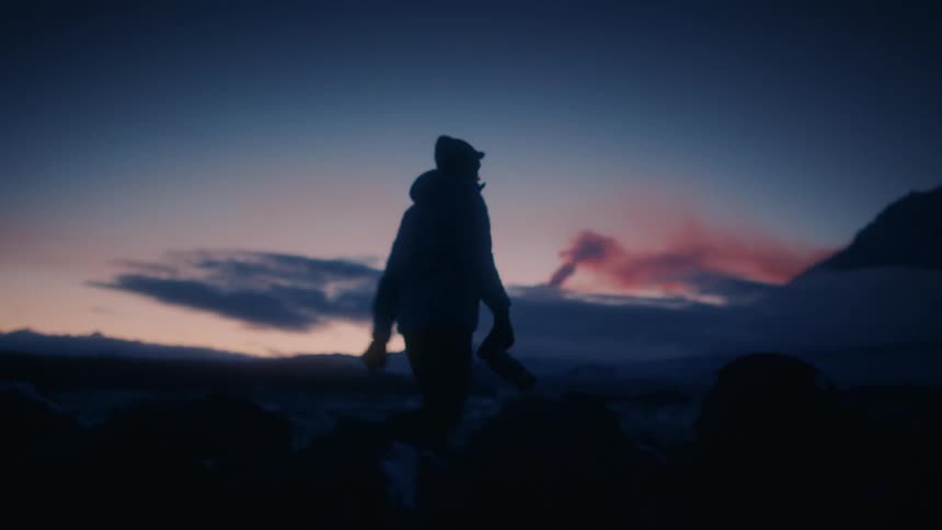 A hiker walks along a rocky trail at dawn with mountains in the distance