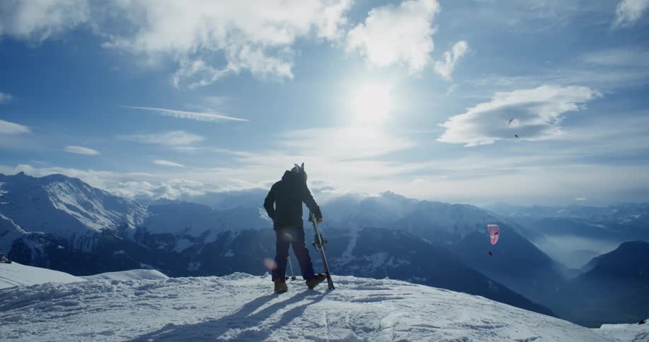 A person standing on the edge of a snowy mountain with their skis while people parasail through the mountains