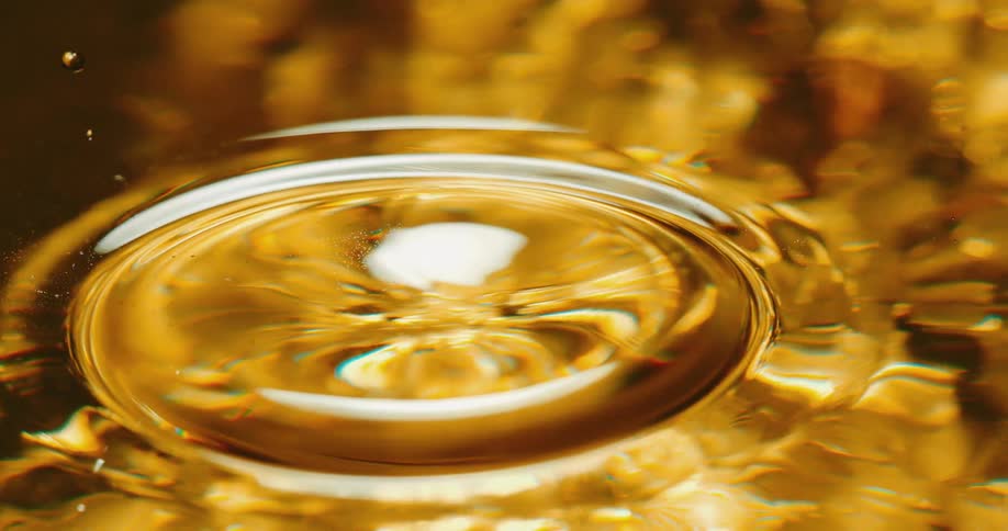 A clear sphere falls into a golden liquid and creates a rippling ring on the surface as it splashes and small droplets fly into the air