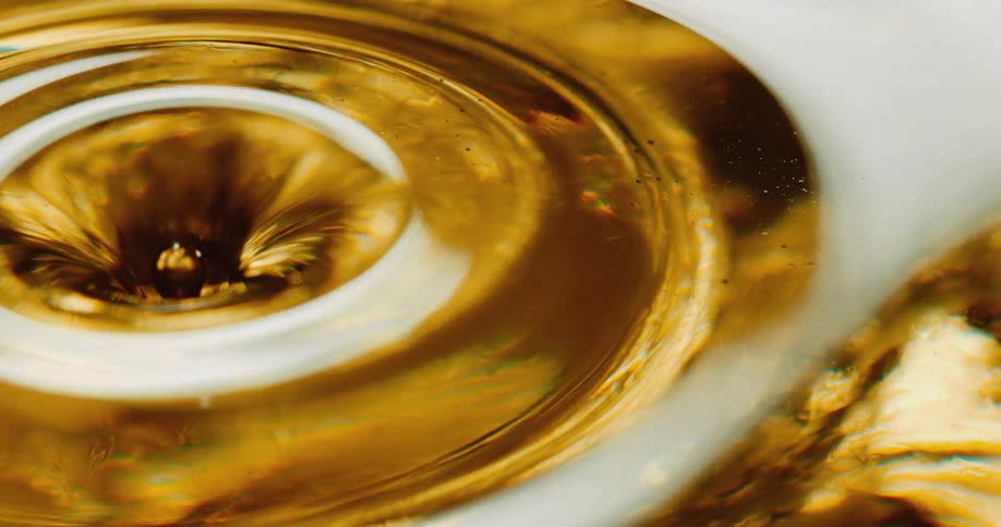 A clear sphere falls into a golden liquid and creates a rippling ring on the surface and splashes back up and down and then another orb falls