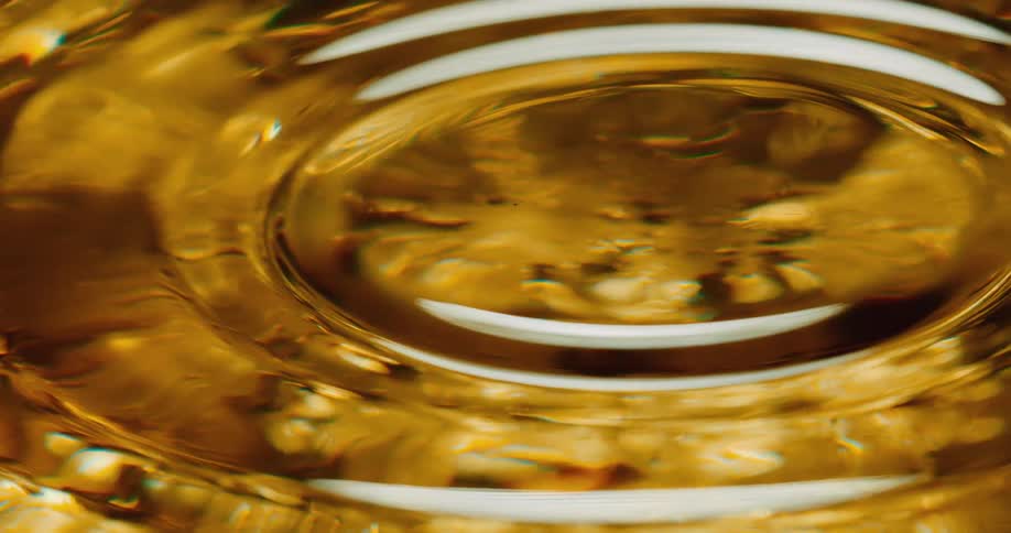 A gold clear liquid with a droplet being dropped onto the smooth surface creating ripples across the surface