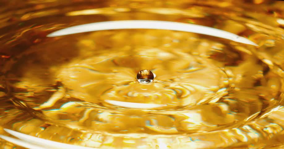 Drop falls into amber fluid forming a brief sphere in middle of expanding concentric ring of ripples