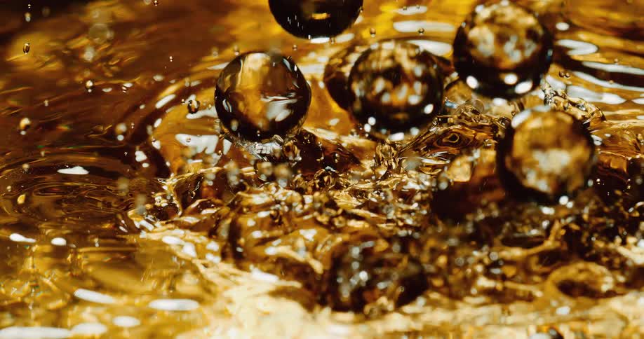 Clear spheres fall into a golden liquid and ripples move across the surface as tiny droplets fly into the air