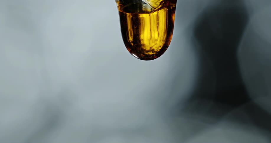 Light shines on a clear golden liquid as it stretches and drips as a small sphere breaks free and falls