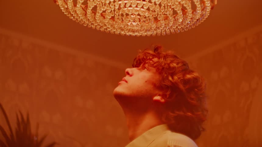 A man standing under a glass chandelier looking up and closing his eyes