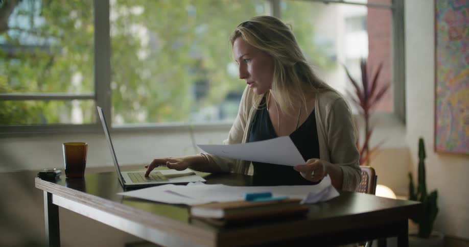 A woman looking at a paper in her hand and typing on a laptop there are also papers and a cup on the table which shows that she is working on a project or assignment