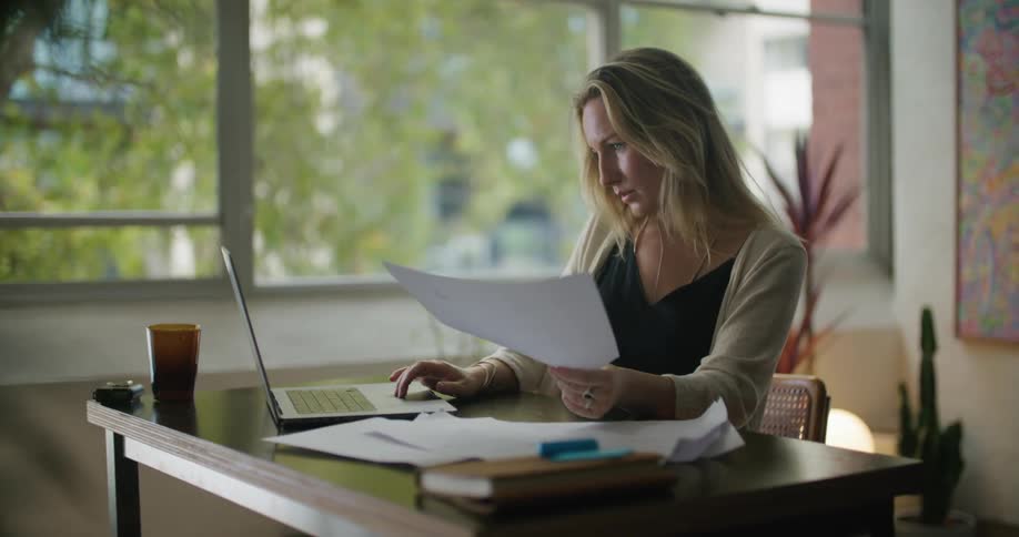 Blonde businesswoman with black top and beige cardigan sits at desk using laptop at the office