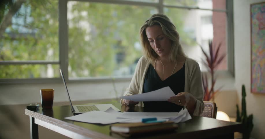 A woman is sitting at a desk with a laptop computer and going through paperwork