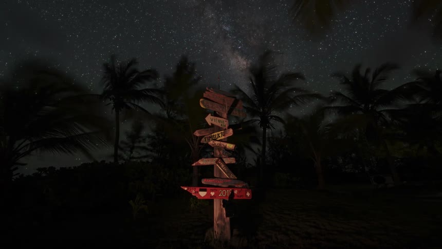 A sign post with signs pointing in many directions stands near palm trees as stars and clouds move across the night sky
