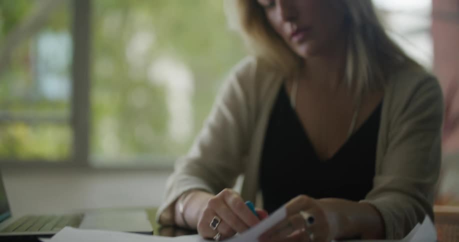 A woman sitting at her desk highlighting papers next to a laptop as she looks contemplatively