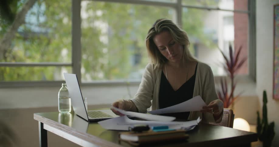 A woman is sitting at a desk with a laptop computer and looking at paperwork