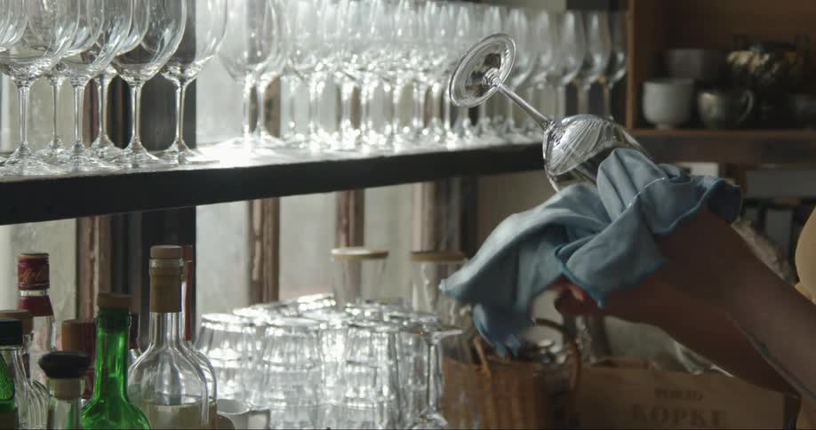 A bartender is taking wine glasses from a shelf and cleaning them with a cloth in a restaurant