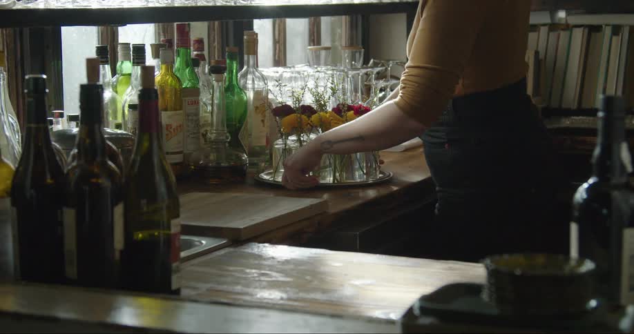 Woman places tray of flowers in bottles on wooden bar counter strewn with glasses and liquor bottles