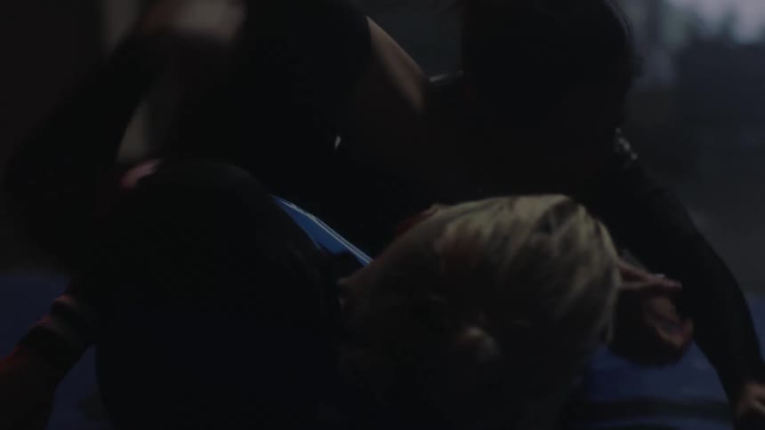 Two women a blonde and a brunette wrestle agressively on a sports mat with the blonde woman holding the other in a tight grip