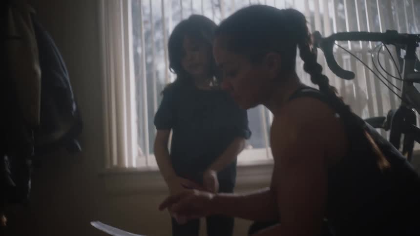 Woman crouches next to bicycle in house near window and examines paper handed to her by her daughter