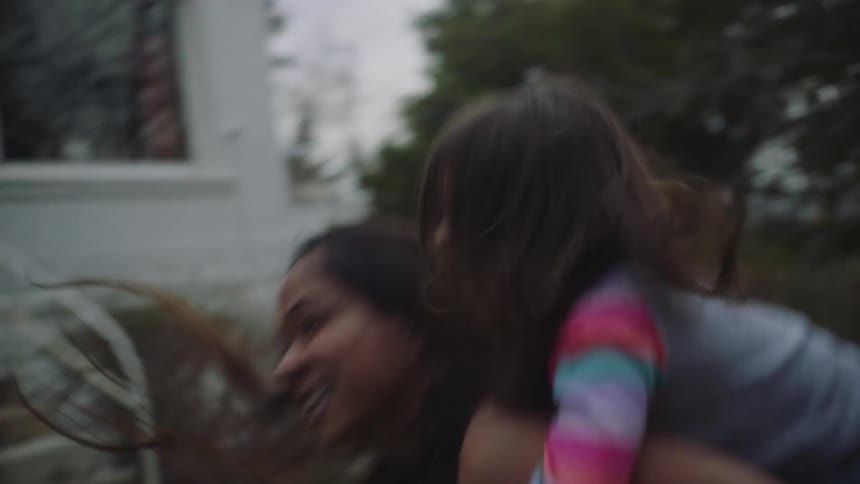 A little girl is riding on her mothers back as they laugh and play in the yard in front of their house