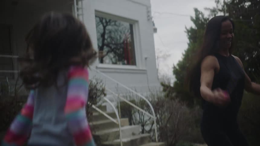 A woman is in front of her house jumping rope as her young daughter watches and jumps imitating her mom