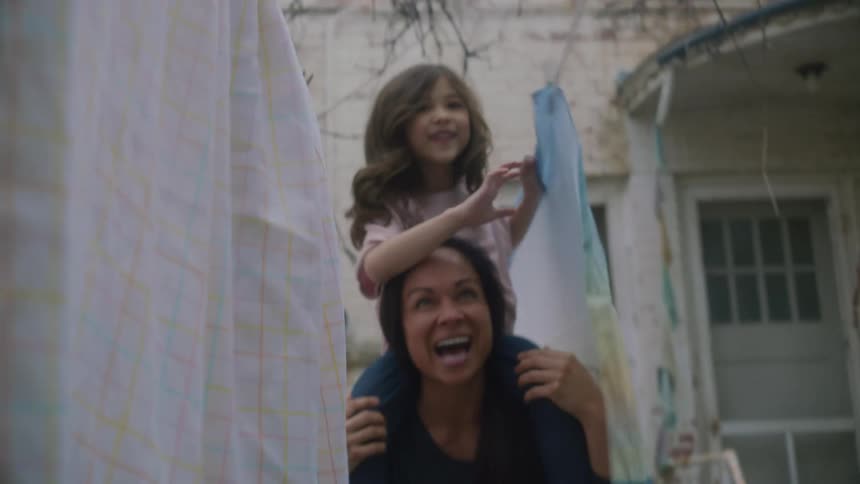 A little girl is sitting on a womans shoulders walking through sheets on a clothes line near a building